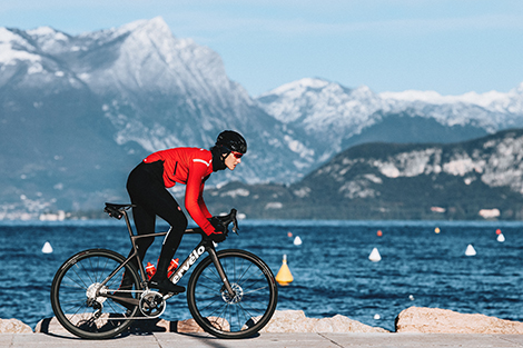 vrouw-in-lange-fietsbroek-en-fietsjas-fietsend-langs-meer-en-bergen-met-sneeuw.jpg